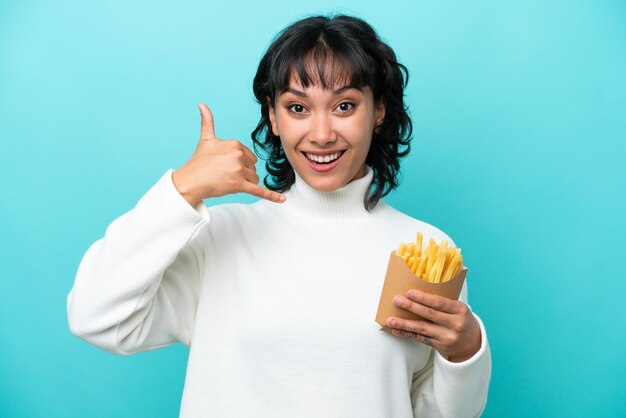 Young Argentinian woman holding fried chips isolated on blue background making phone gesture Call me back sign