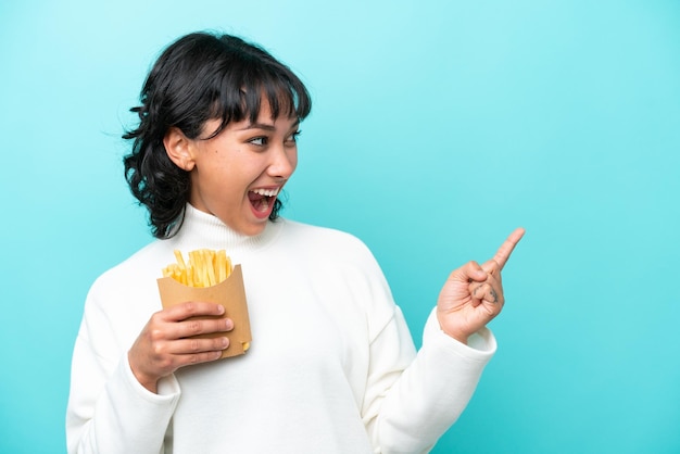 Young Argentinian woman holding fried chips isolated on blue background pointing finger to the side and presenting a product