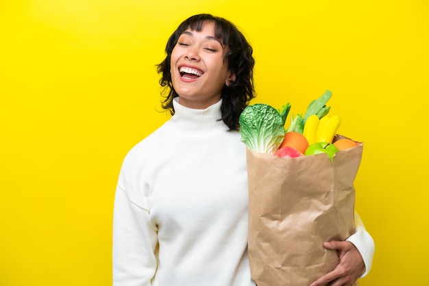 Young argentinian woman holding a grocery shopping bag isolated on yellow background laughing