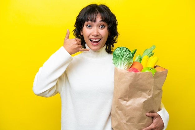 노란색 배경에 격리된 식료품 쇼핑백을 들고 전화 제스처를 취하는 젊은 아르헨티나 여성