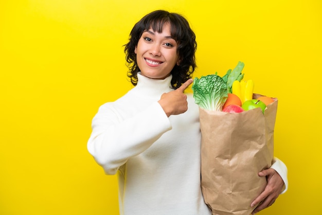 노란색 배경에 격리된 식료품 쇼핑백을 들고 뒤를 가리키는 젊은 아르헨티나 여성
