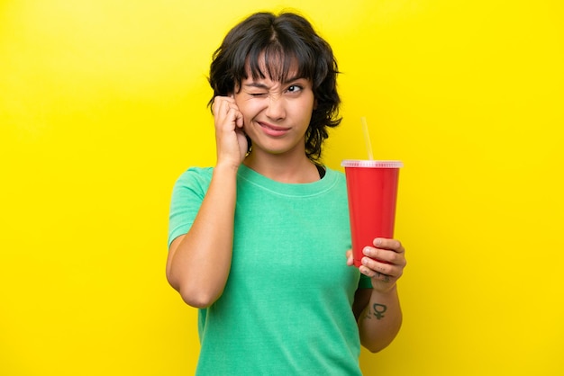 Young Argentinian woman holding a soda isolated on yellow background frustrated and covering ears