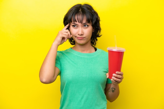 Young Argentinian woman holding a soda isolated on yellow background having doubts and thinking