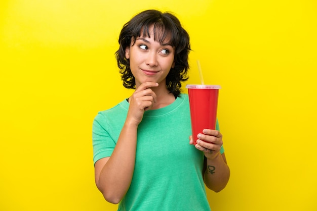 Young Argentinian woman holding a soda isolated on yellow background looking up while smiling