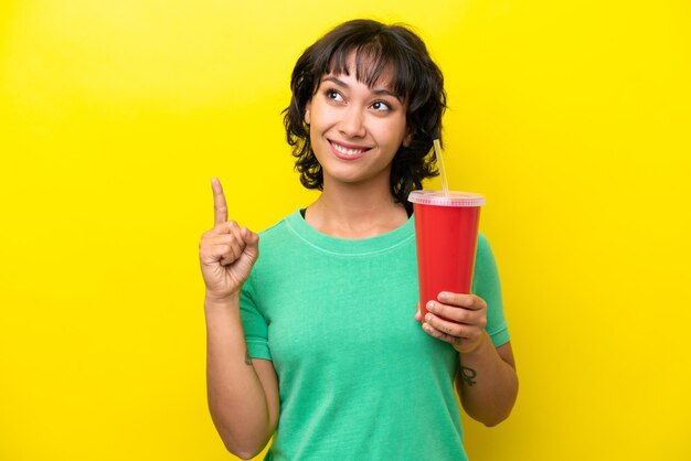 Young argentinian woman holding a soda isolated on yellow background pointing up a great idea