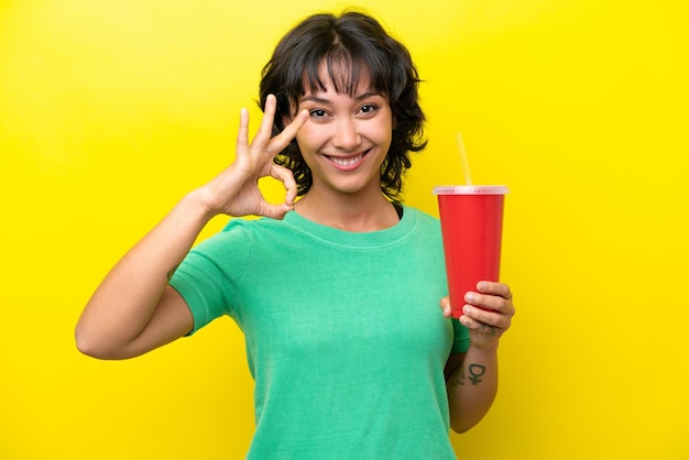 Young argentinian woman holding a soda isolated on yellow background showing ok sign with fingers