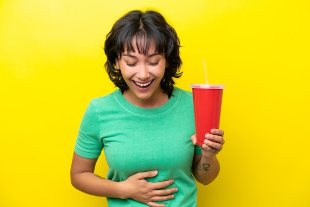 Young argentinian woman holding a soda isolated on yellow background smiling a lot