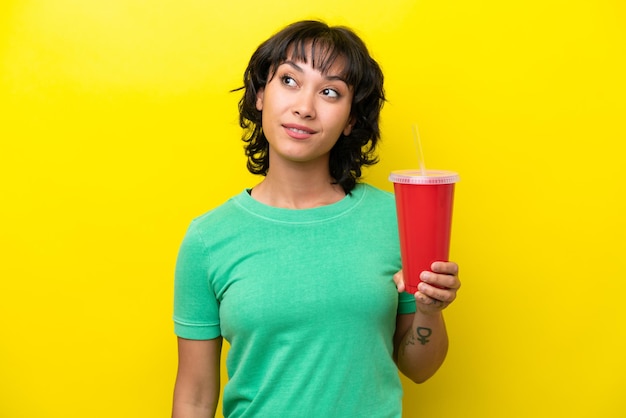 Young Argentinian woman holding a soda isolated on yellow background thinking an idea while looking up