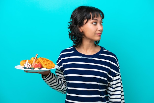 Young Argentinian woman holding waffles isolated on blue background looking to the side