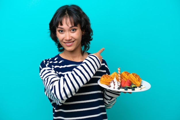 Young Argentinian woman holding waffles isolated on blue background pointing back