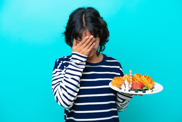 피곤하고 아픈 표정으로 파란색 배경에 고립된 와플을 들고 있는 젊은 아르헨티나 여성