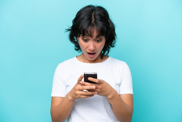 Молодая аргентинская женщина, изолированная на синем фоне, смотрит в камеру, используя мобильный телефон, с удивленным выражением лица