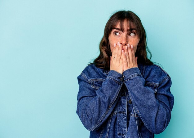 Photo young argentinian woman isolated on blue background thoughtful looking to a copy space covering mouth with hand.