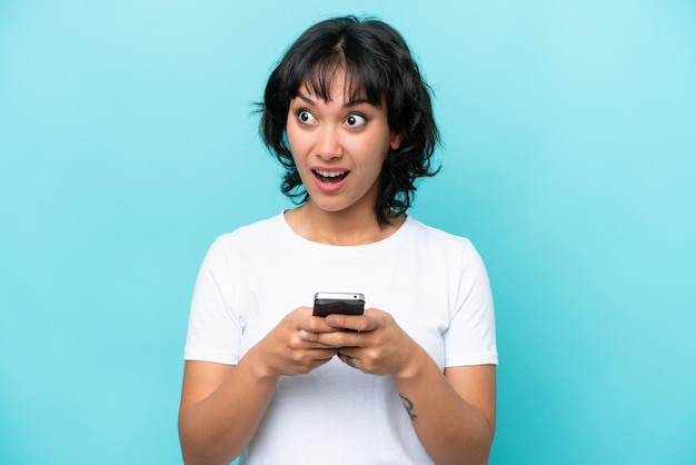 Young Argentinian woman isolated on blue background using mobile phone and looking up