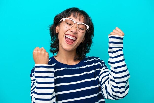 Young Argentinian woman isolated on blue background With glasses and celebrating a victory