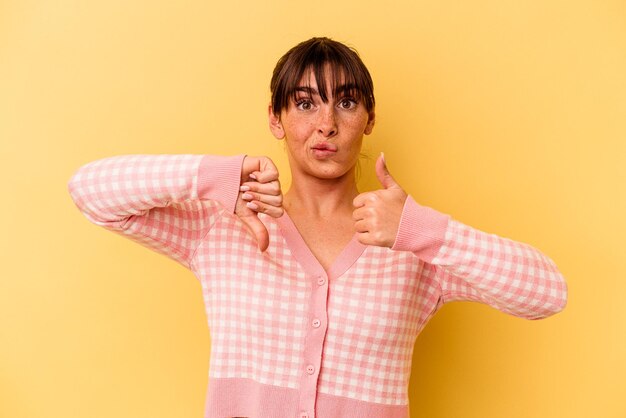 Young Argentinian woman isolated on yellow background showing thumbs up and thumbs down difficult choose concept