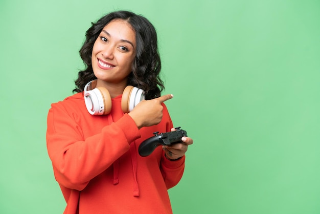 Young Argentinian woman playing with a video game controller over isolated background pointing back