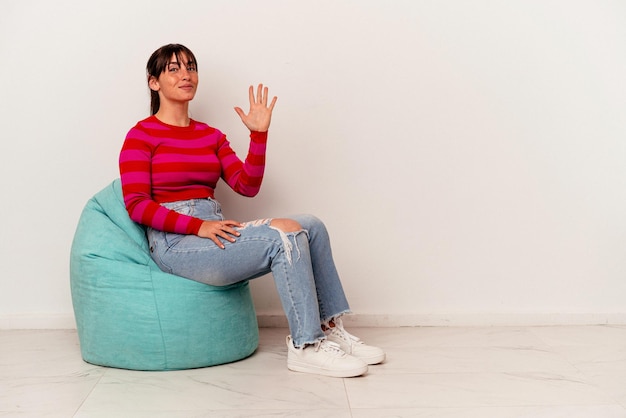 Young Argentinian woman sitting on a puff isolated on white background smiling cheerful showing number five with fingers.