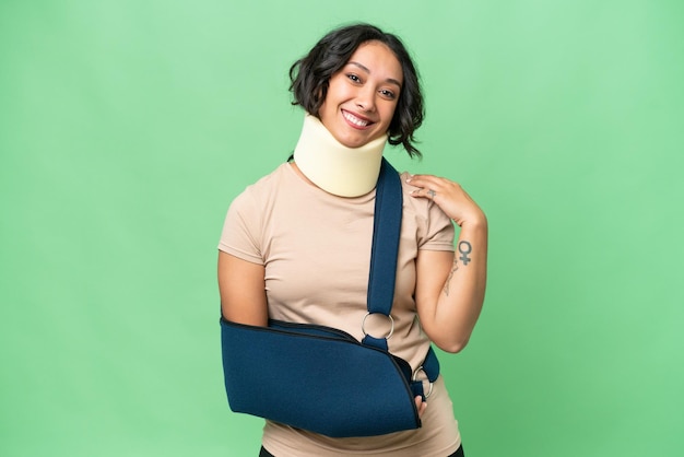 Young argentinian woman wearing neck brace and sling over isolated background laughing