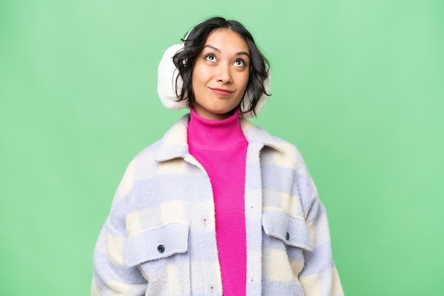 Young Argentinian woman wearing winter muffs over isolated background and looking up