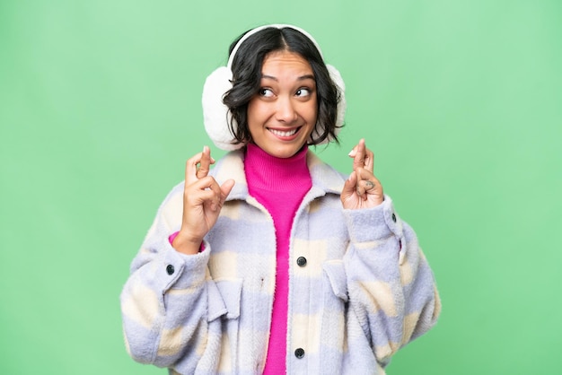 Young argentinian woman wearing winter muffs over isolated background with fingers crossing