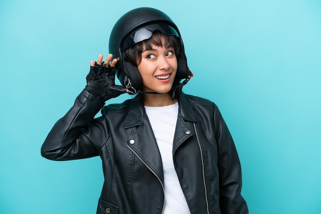 Young Argentinian woman with a motorcycle helmet isolated on blue background listening to something by putting hand on the ear