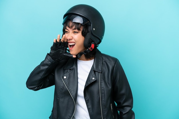 Young argentinian woman with a motorcycle helmet isolated on\
blue background shouting with mouth wide open to the side