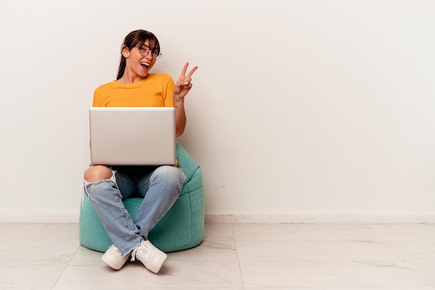 Young Argentinian woman working with pc sitting on a puff isolated on white background joyful and carefree showing a peace symbol with fingers.