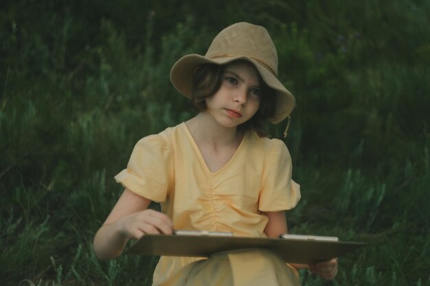 young artist 1013 years old in a yellow dress and a straw hat in nature painting a picture