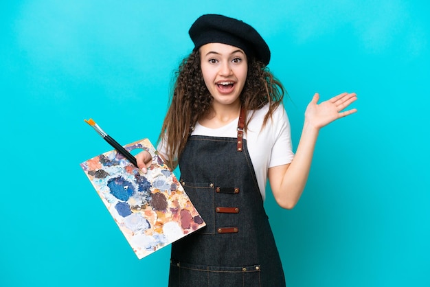 Молодая художница-арабка, держащая палитру на синем фоне с потрясенным выражением лица