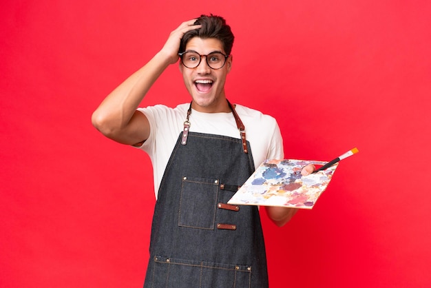 Young artist caucasian man holding a palette isolated on red background with surprise expression