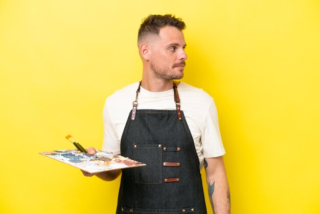 Young artist caucasian man holding a palette isolated on yellow background looking to the side