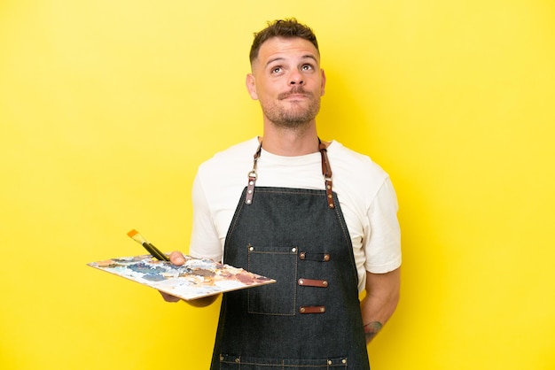 Young artist caucasian man holding a palette isolated on yellow background and looking up