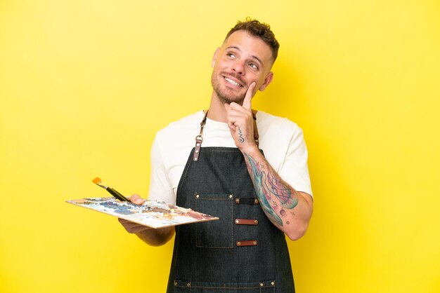 Young artist caucasian man holding a palette isolated on yellow background thinking an idea while looking up