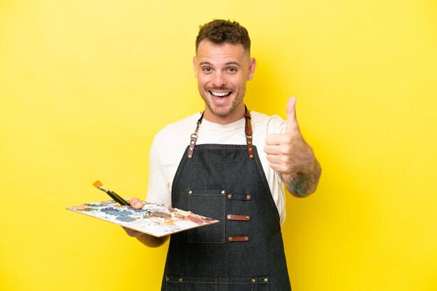 Young artist caucasian man holding a palette isolated on yellow background with thumbs up because something good has happened