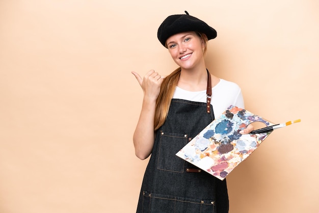 Young artist caucasian woman holding a palette isolated on beige background pointing to the side to present a product