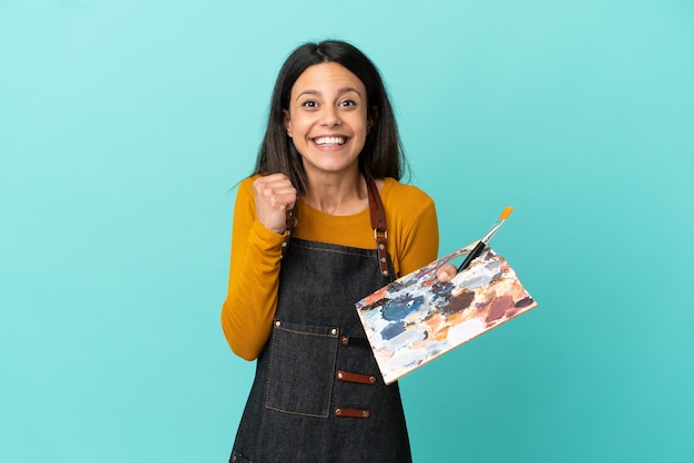 Young artist caucasian woman holding a palette isolated on blue background celebrating a victory in winner position