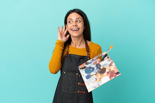 Young artist caucasian woman holding a palette isolated on blue background listening to something by putting hand on the ear