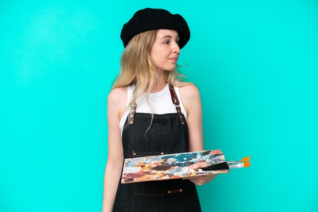 Young artist caucasian woman holding a palette isolated on blue background looking to the side