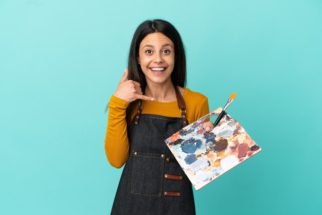 Young artist caucasian woman holding a palette isolated on blue background making phone gesture. Call me back sign