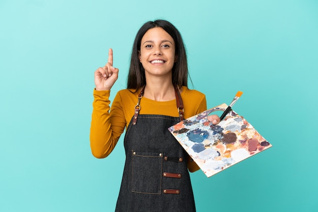 Young artist caucasian woman holding a palette isolated on blue background showing and lifting a finger in sign of the best