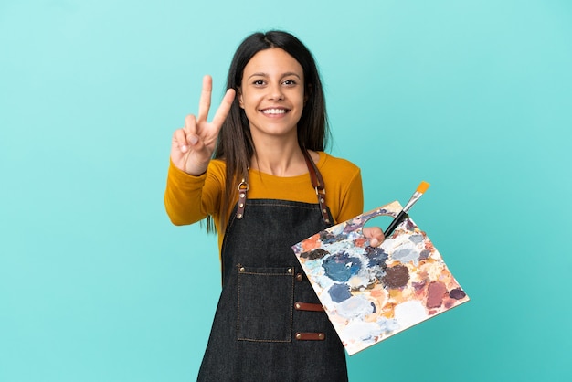 Young artist caucasian woman holding a palette isolated on blue background smiling and showing victory sign