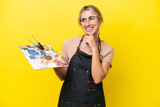 Young artist caucasian woman holding a palette isolated on yellow background looking to the side and smiling