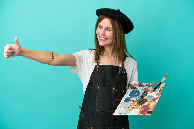 Young artist English woman holding a palette isolated on blue background giving a thumbs up gesture