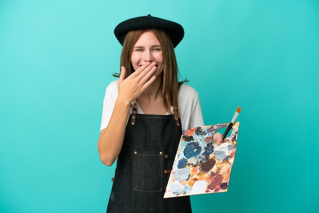 Young artist English woman holding a palette isolated on blue background happy and smiling covering mouth with hand