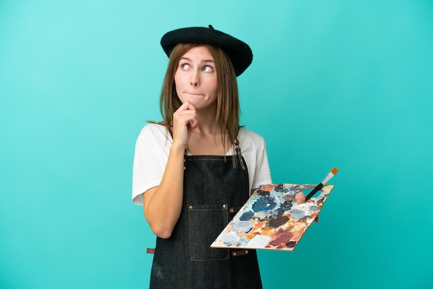 Young artist English woman holding a palette isolated on blue background having doubts and thinking