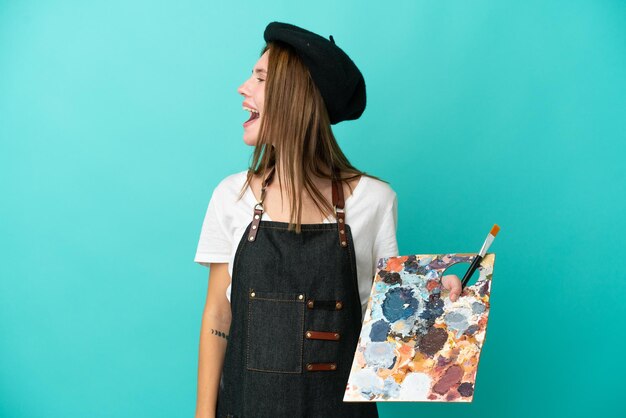 Young artist English woman holding a palette isolated on blue background laughing in lateral position