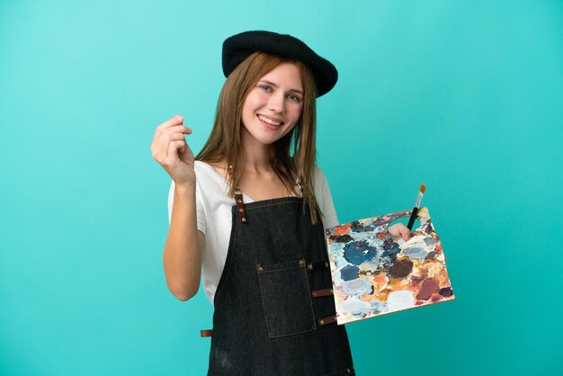 Young artist English woman holding a palette isolated on blue background making money gesture