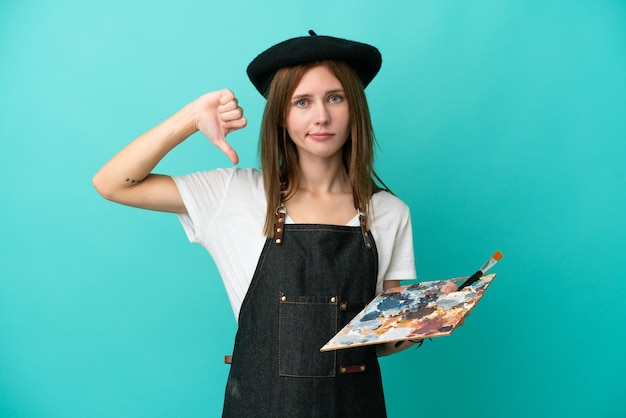 Young artist English woman holding a palette isolated on blue background showing thumb down with negative expression