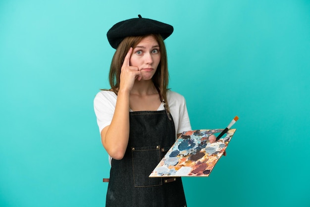 Young artist English woman holding a palette isolated on blue background thinking an idea